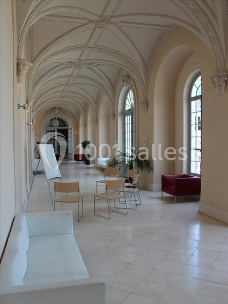 Un couloir lumineux avec voûtes, grandes fenêtres, chaises, un tableau blanc et des plantes décoratives.