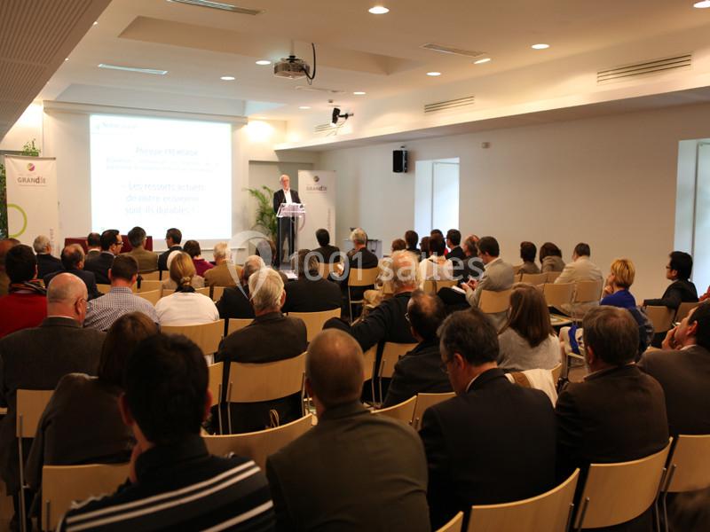 Un conférencier s'adresse à un public assis dans une salle lumineuse équipée d'un projecteur.