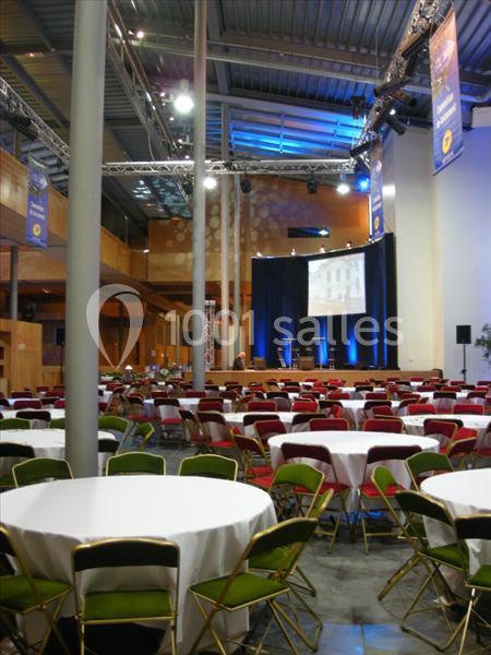 Salle de conférence avec tables rondes dressées, chaises colorées et une scène équipée d'un écran et de projecteurs.