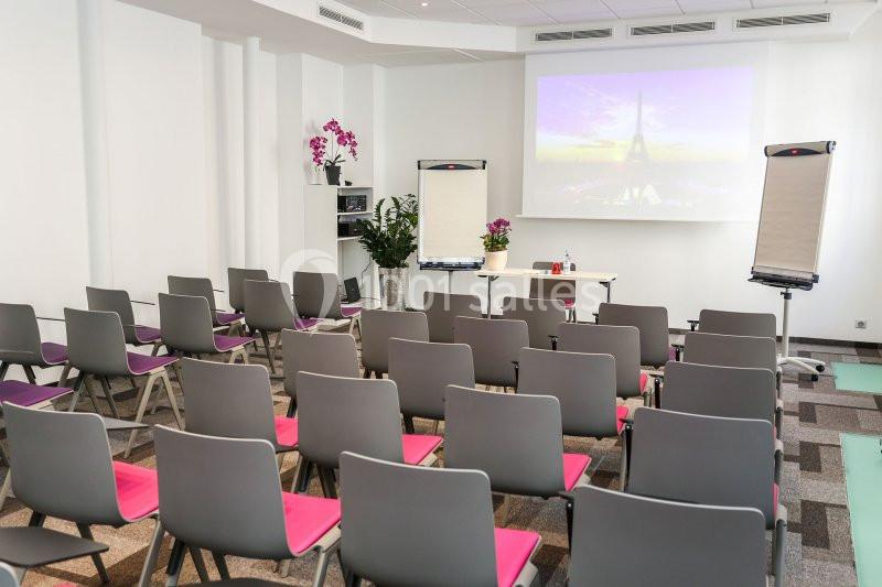 Salle de conférence lumineuse avec chaises grises, écrans de projection, tableaux blancs et décorations florales.
