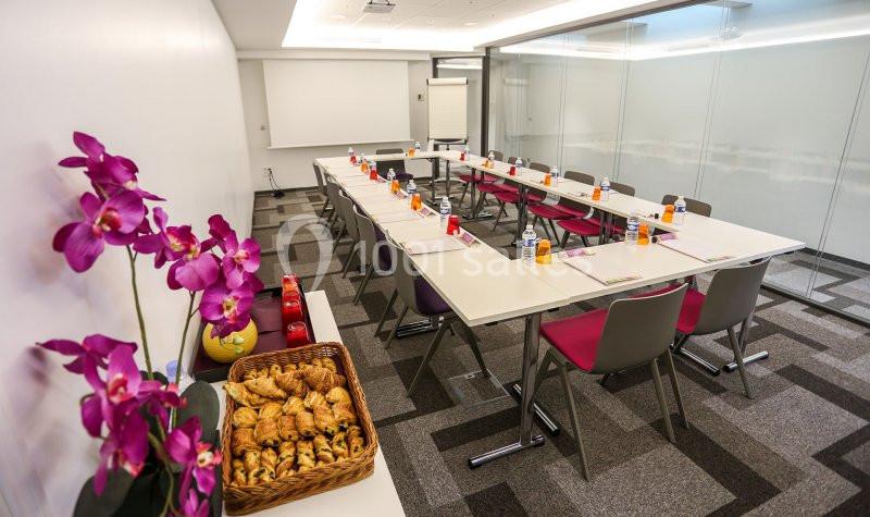 Salle de réunion lumineuse avec tables en U, chaises colorées, viennoiseries et boissons disposées sur une table.