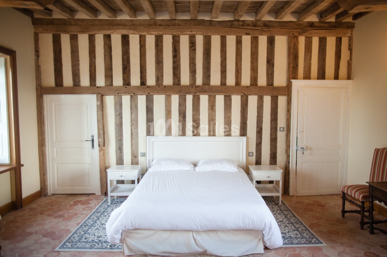 Chambre avec lit double, murs à colombages, sol en terre cuite, tapis et mobilier blanc.