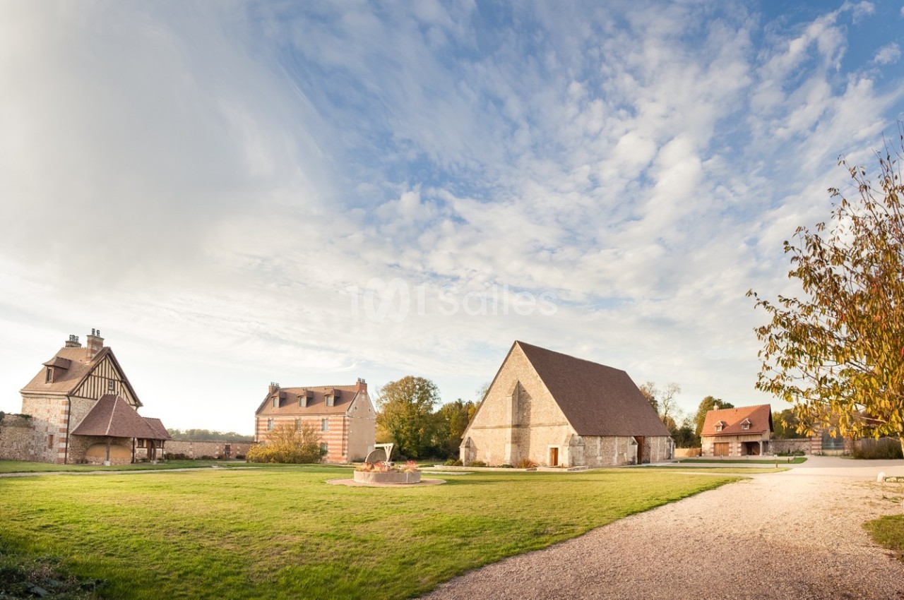 Vue d'un domaine rural avec bâtiments anciens en pierre, pelouse, chemin de gravier et ciel partiellement nuageux.
