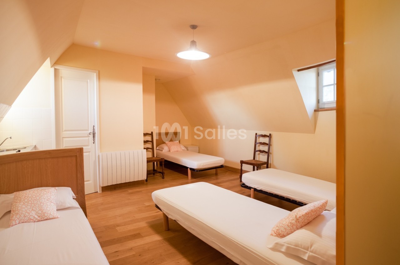 Chambre mansardée avec trois lits simples, parquet en bois, murs clairs et une petite fenêtre apportant de la lumière…