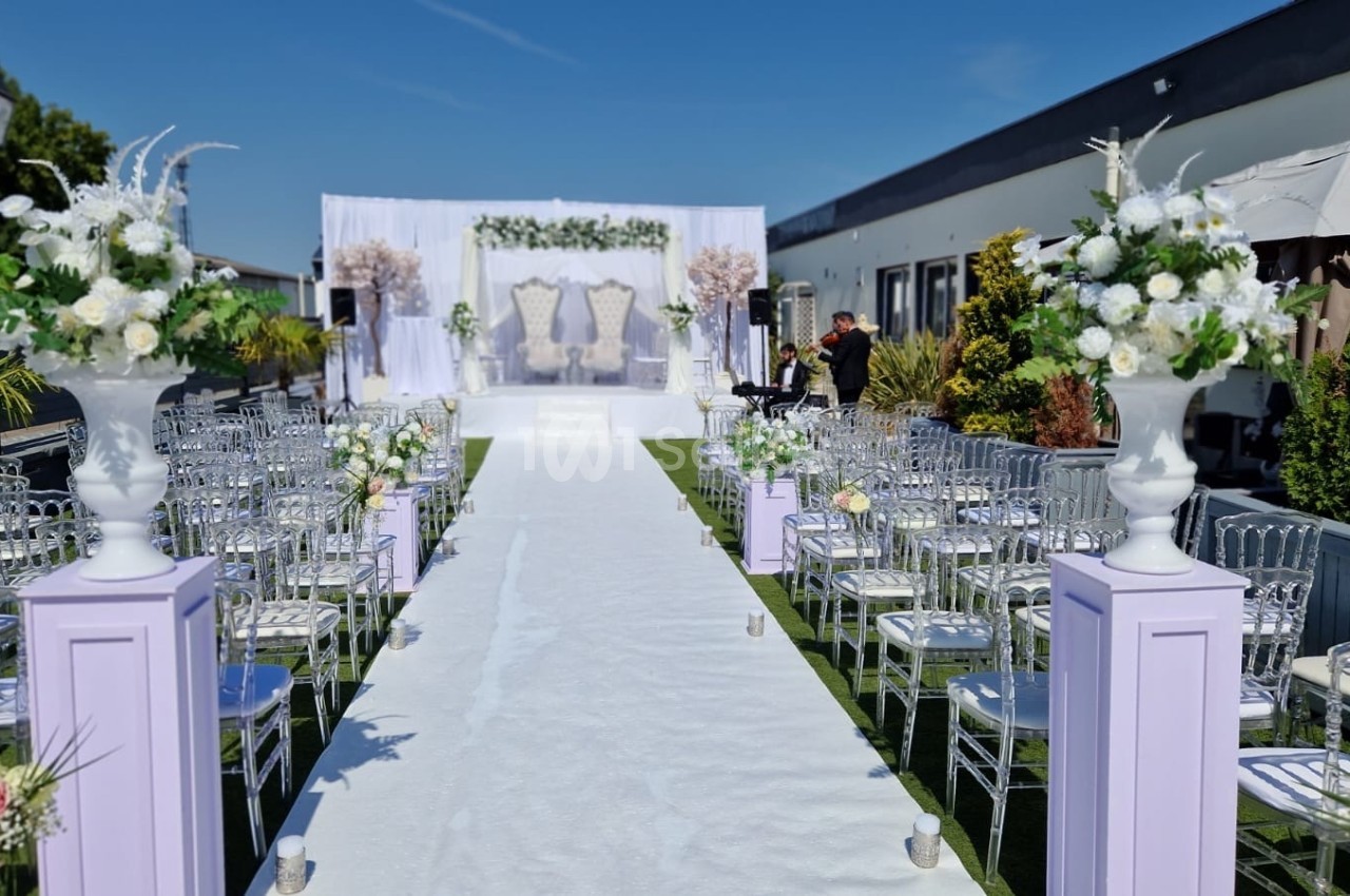 Allée centrale décorée pour une cérémonie en plein air, avec chaises transparentes et fleurs blanches.