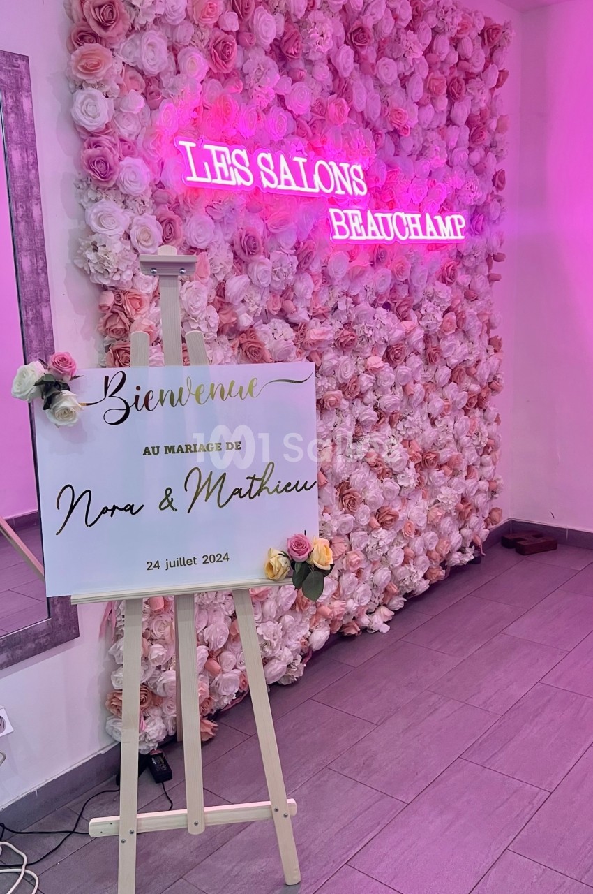 Mur de fleurs roses avec un panneau sur chevalet annonçant un mariage, éclairage néon en arrière-plan.