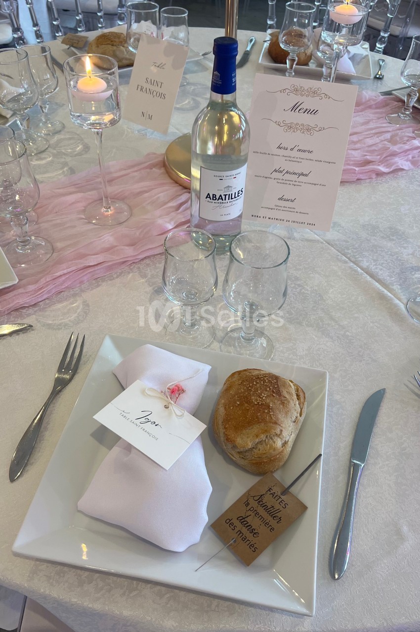Table dressée avec une assiette carrée, serviette pliée, pain, menu, bougie, et bouteille d'eau minérale.