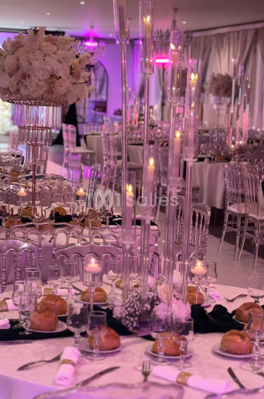 Salle de réception élégante avec tables dressées, chandeliers en verre, bougies allumées et décoration florale blanche.