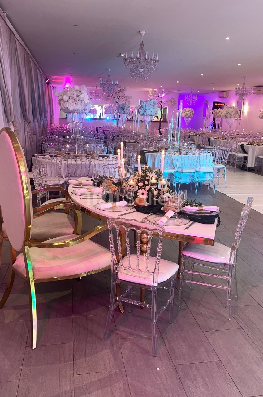 Salle de réception décorée avec des tables élégantes, des chandeliers et des chaises transparentes sous un éclairage coloré.