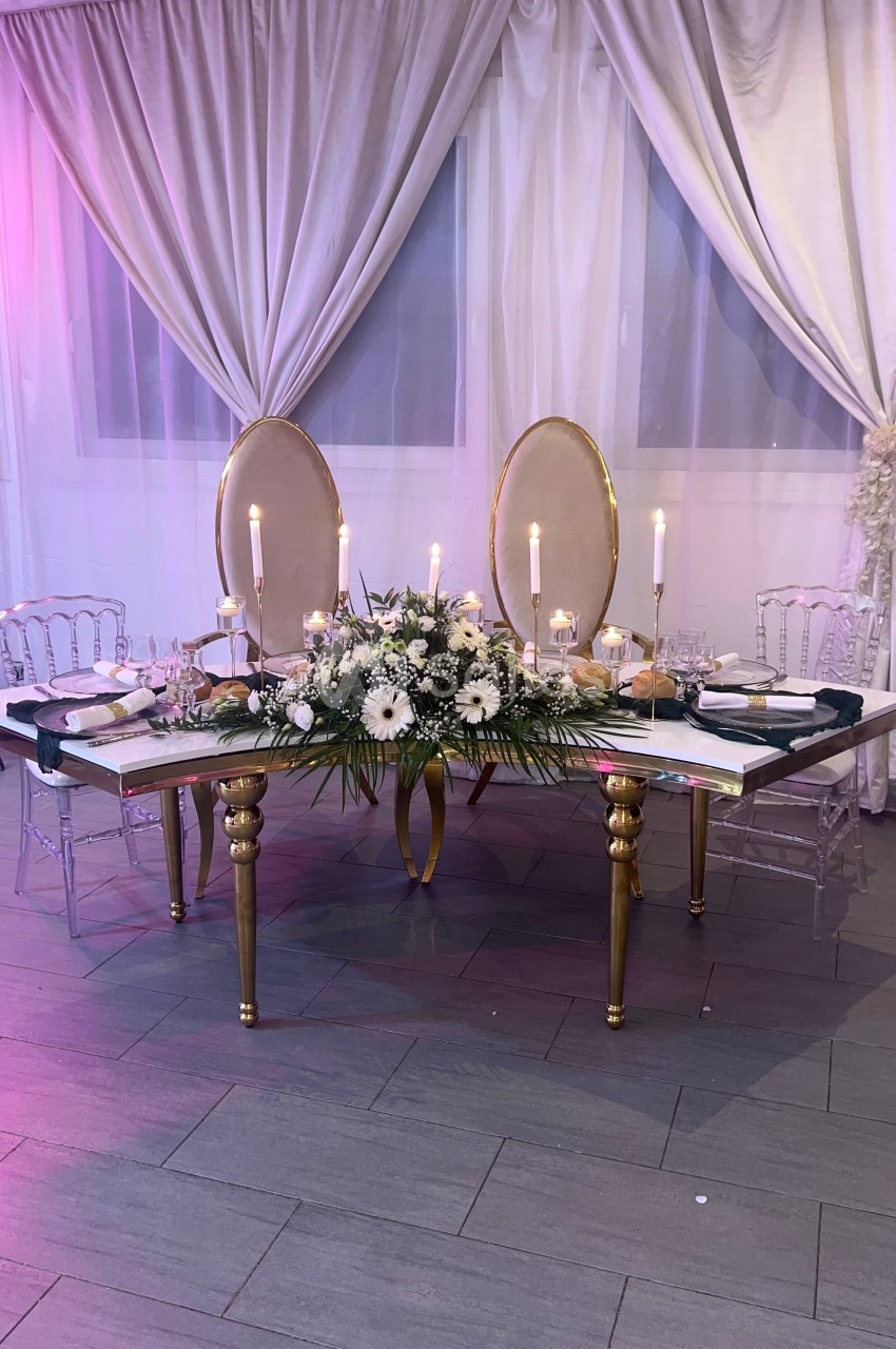 Table élégante décorée de fleurs blanches, bougies allumées et chaises transparentes, devant des rideaux clairs.