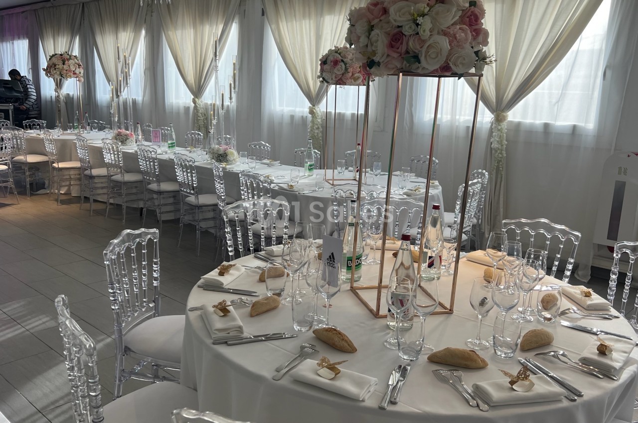 Salle de réception décorée avec des tables rondes dressées, chaises transparentes et centres de table fleuris.