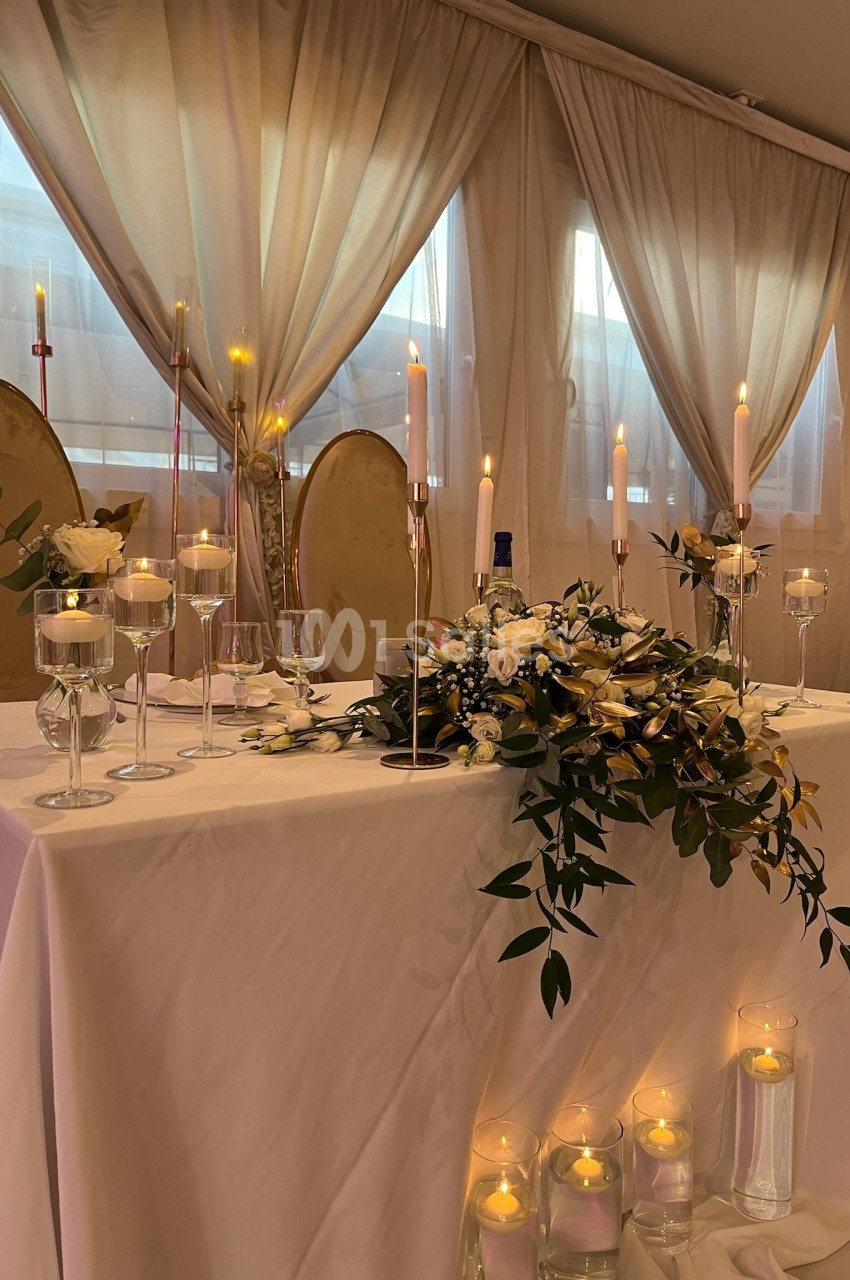 Table décorée pour un mariage avec nappes blanches, bougies allumées, fleurs et chaises dorées en arrière-plan.