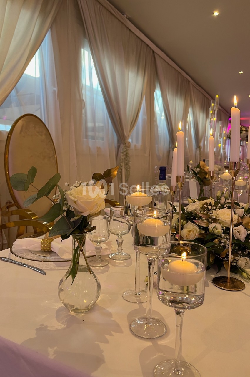 Table élégamment décorée avec des bougies allumées, des fleurs blanches et des verres, dans une salle aux rideaux clairs.