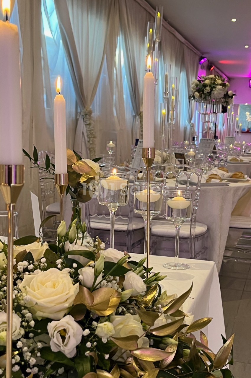 Salle de réception élégante avec tables décorées de fleurs blanches, bougies allumées et chaises transparentes.