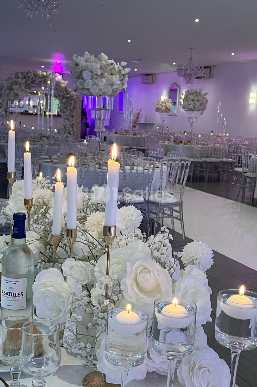 Salle de réception élégante décorée de fleurs blanches, chandeliers et bougies allumées sur des tables dressées.