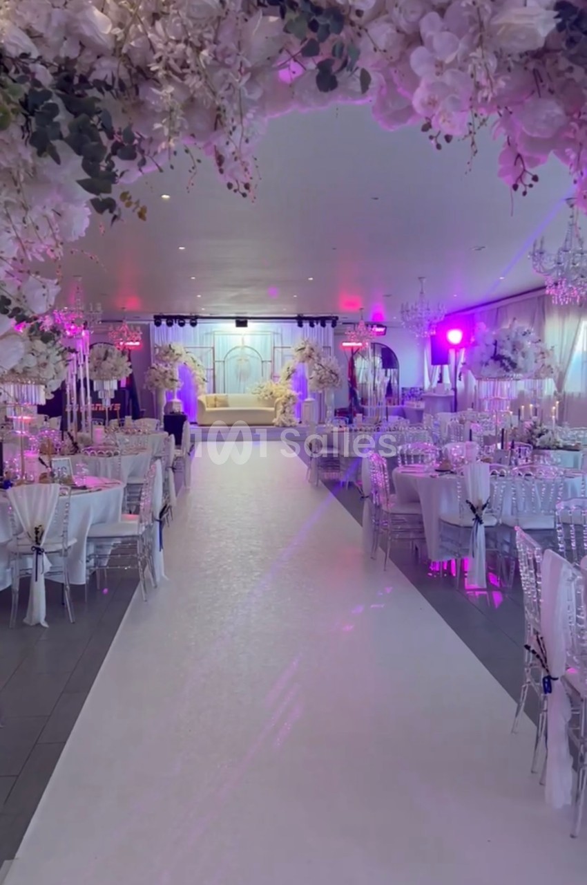 Salle de réception décorée avec des fleurs blanches, tables dressées et éclairage rose, allée centrale menant à une scène.