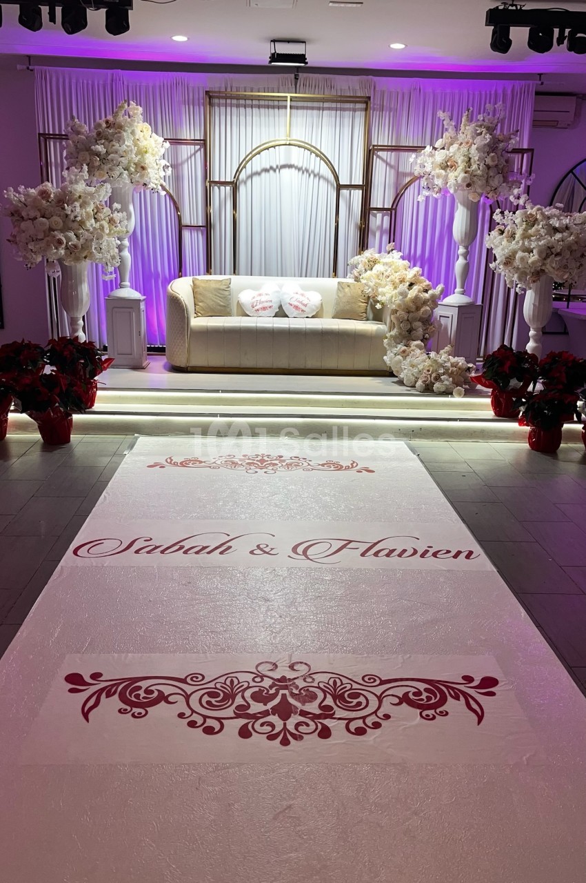 Décor de mariage avec canapé beige, fleurs blanches et tapis personnalisé portant des prénoms en rouge.