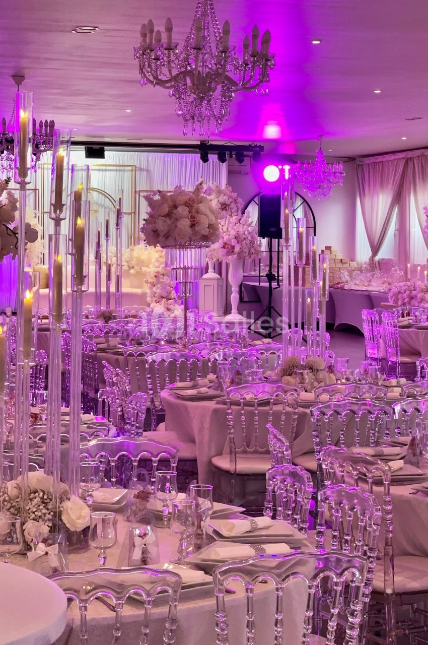 Salle de réception décorée avec des chandeliers, des fleurs blanches et des chaises transparentes sous un éclairage rose.