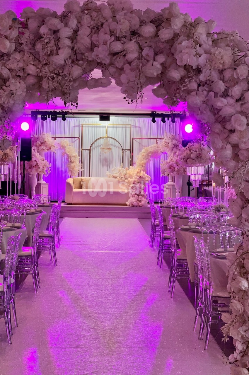 Salle de réception décorée avec des arches fleuries, des tables dressées et un éclairage rose.