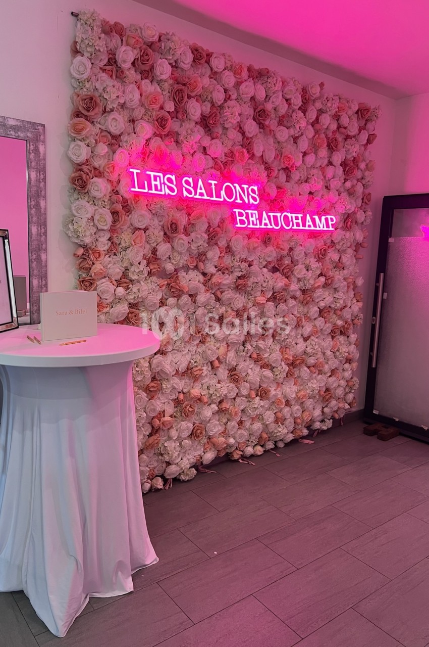 Mur de fleurs roses et blanches avec une enseigne néon rose ’Les Salons Beauchamp’, table haute drapée à gauche.