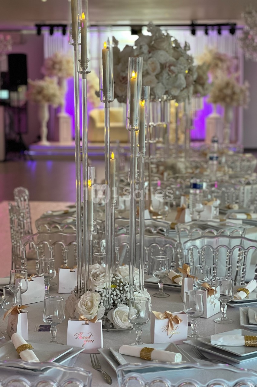 Table de réception élégamment décorée avec chandeliers, fleurs blanches et détails dorés dans une salle lumineuse.