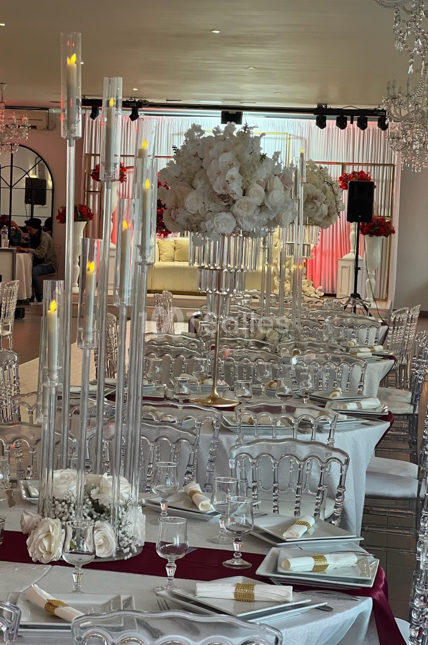 Salle de réception élégante avec tables dressées, chaises transparentes et décorations florales blanches et rouges.