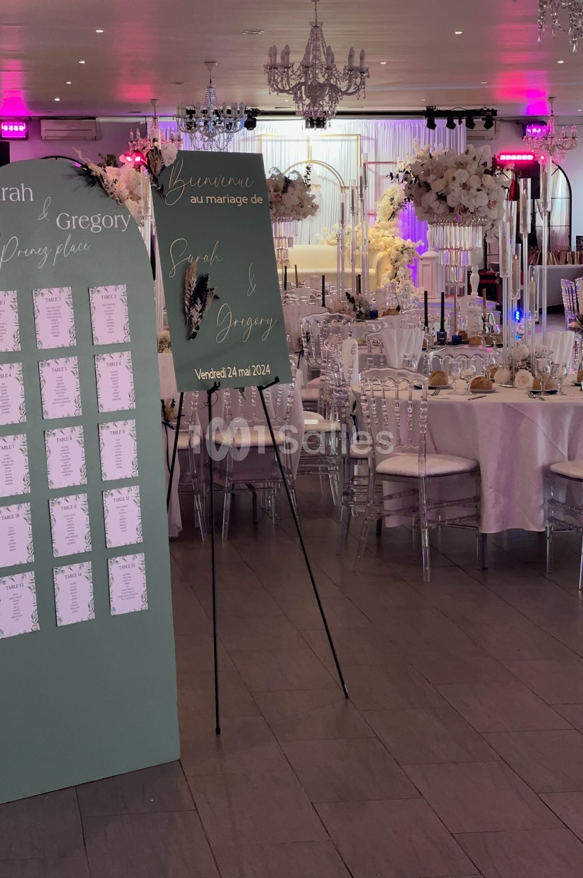 Salle de réception décorée pour un mariage avec panneaux de bienvenue et plan de table, tables dressées et fleurs blanches.