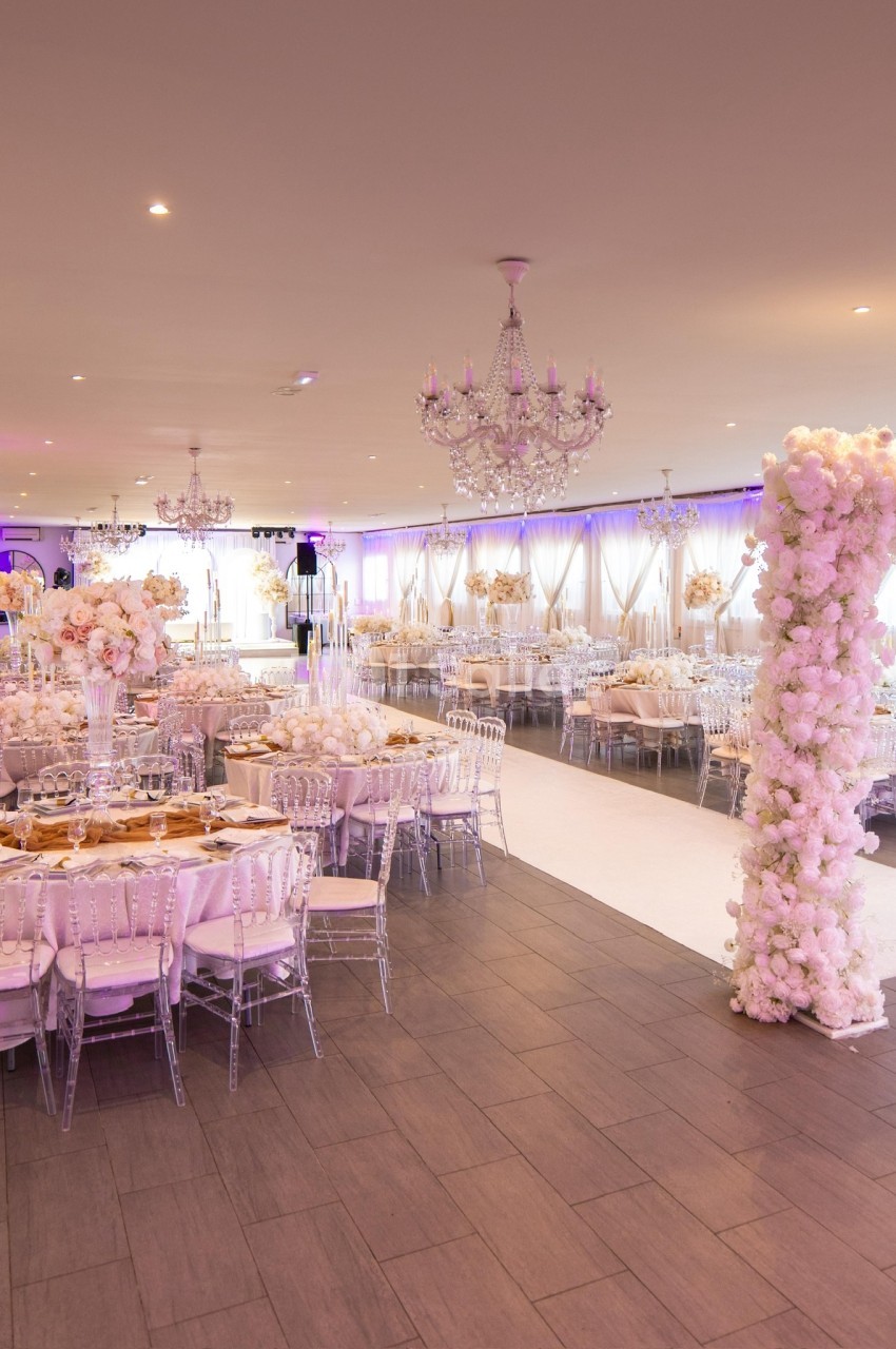 Salle de réception élégante décorée de fleurs blanches, avec tables dressées et chaises transparentes.