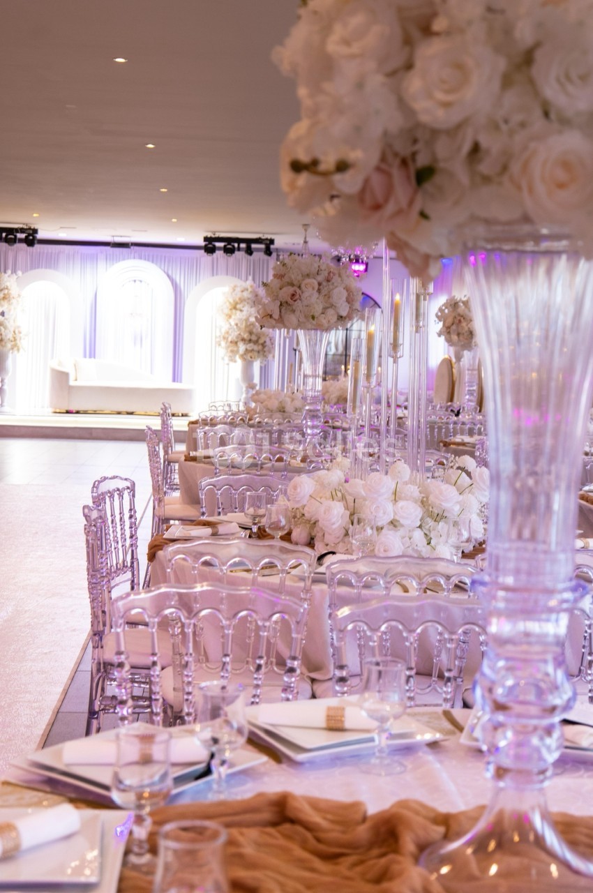 Salle de réception élégante avec tables décorées de fleurs blanches, chaises transparentes et éclairage tamisé.