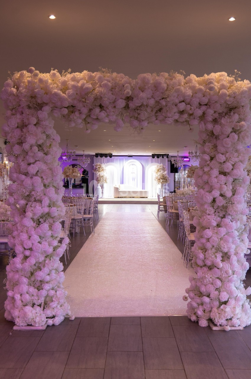Arche florale blanche encadrant une allée menant à une scène décorée pour un événement festif en intérieur.