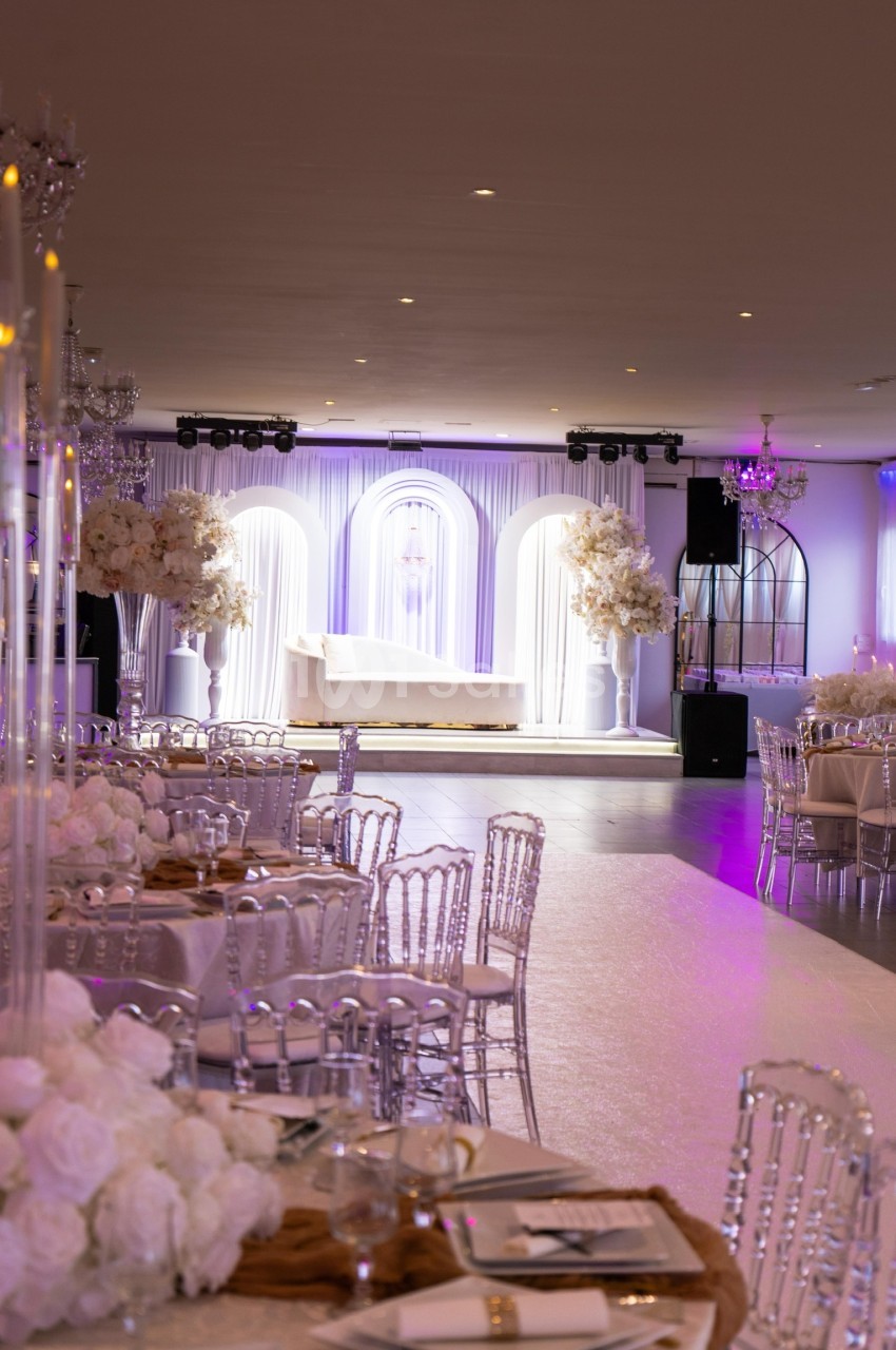 Salle de réception élégante avec tables décorées, chaises transparentes et éclairage violet, devant une scène ornée d'arches.