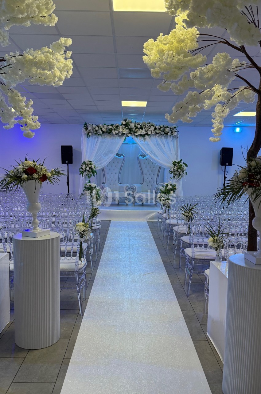 Allée centrale décorée de fleurs blanches menant à une arche ornée pour une cérémonie dans une salle lumineuse.