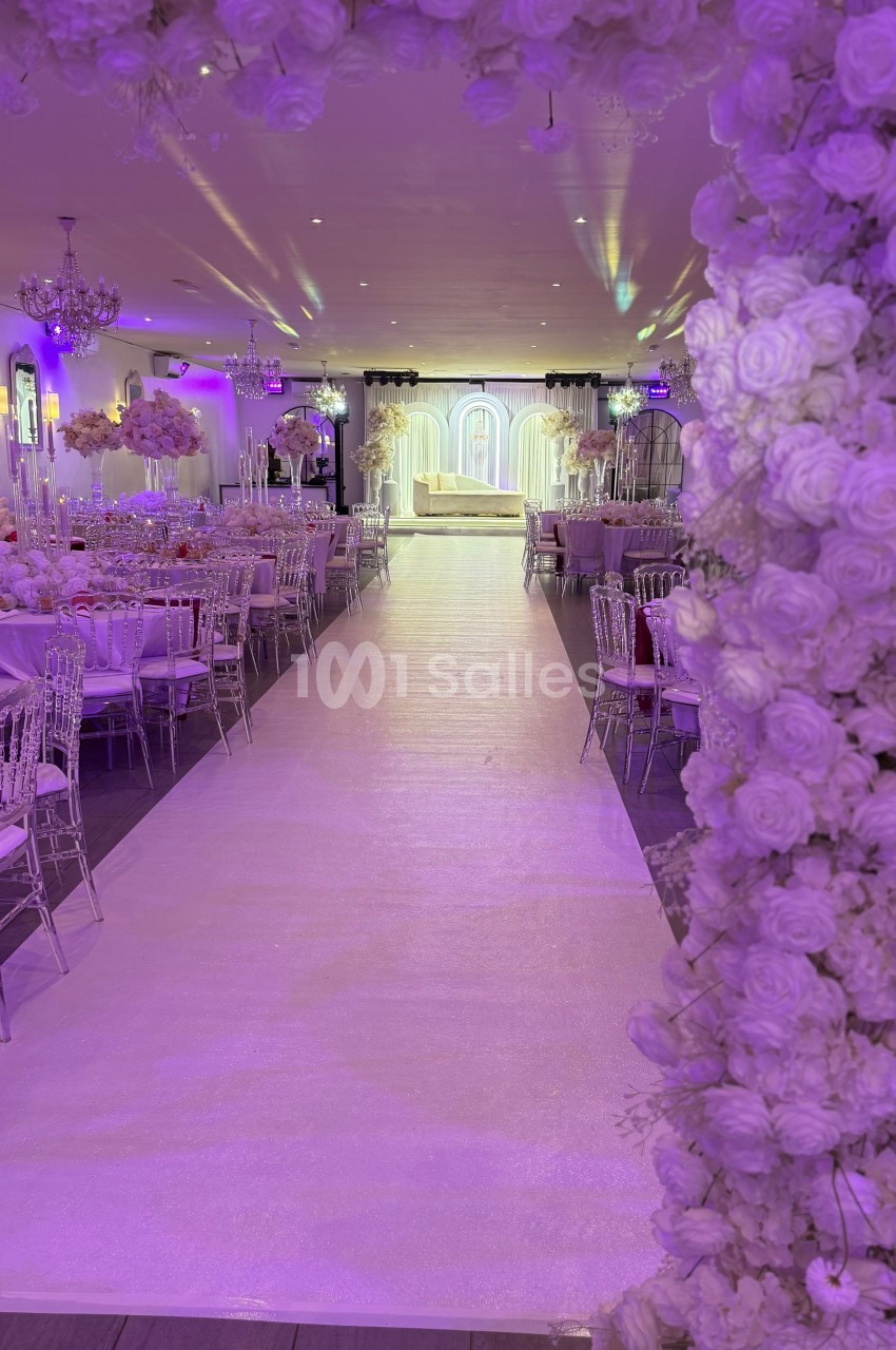 Allée centrale décorée de fleurs blanches menant à une scène illuminée dans une salle de réception élégante.