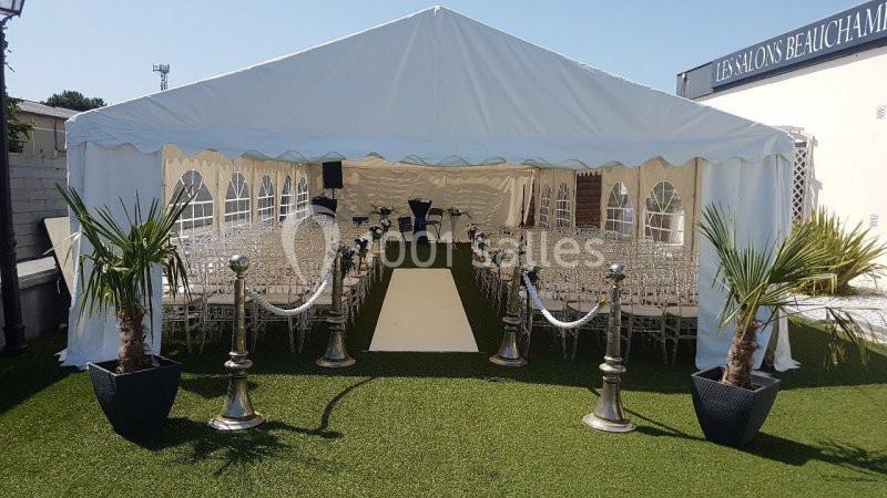 Tente blanche aménagée avec des rangées de chaises transparentes, allée centrale beige et décorations végétales sur gazon.