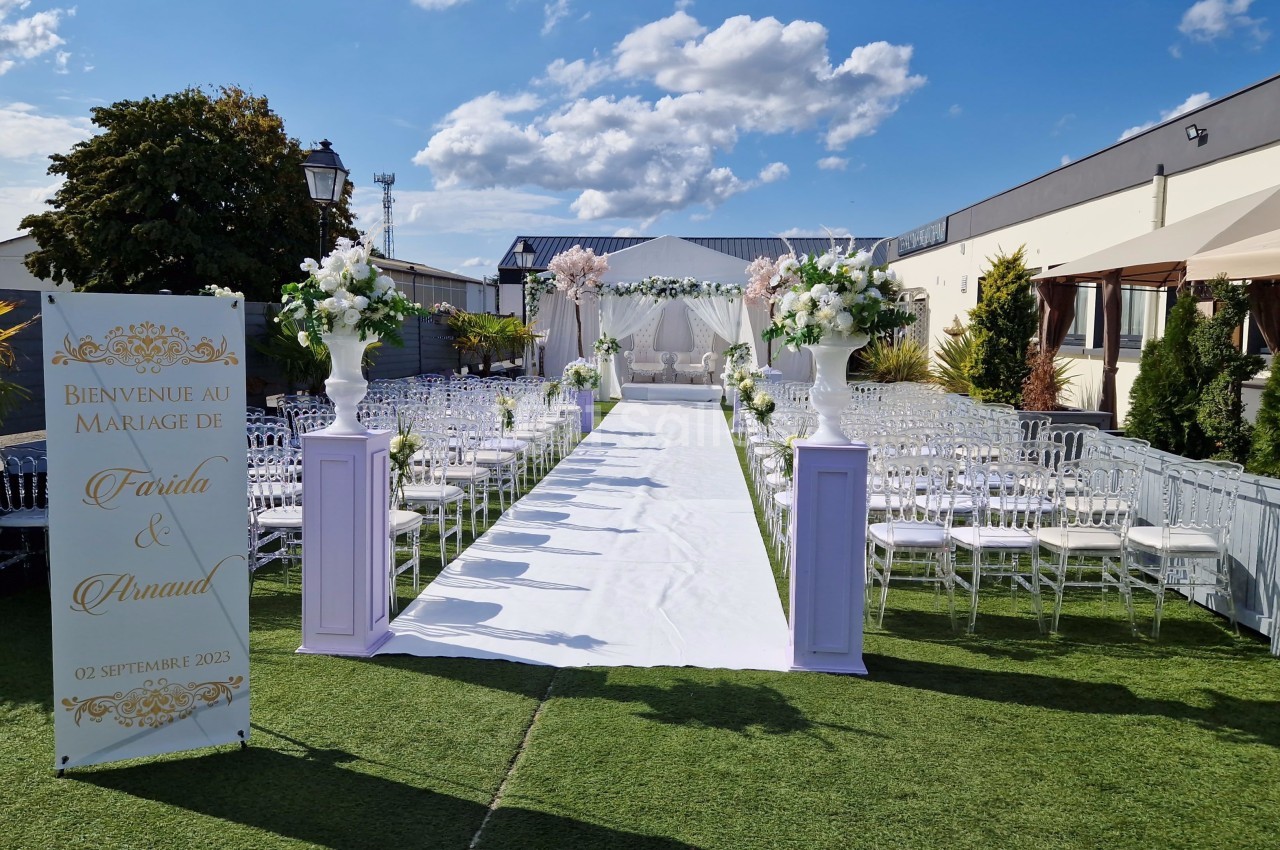 Allée décorée pour une cérémonie de mariage en extérieur, avec chaises transparentes et arche florale au fond.