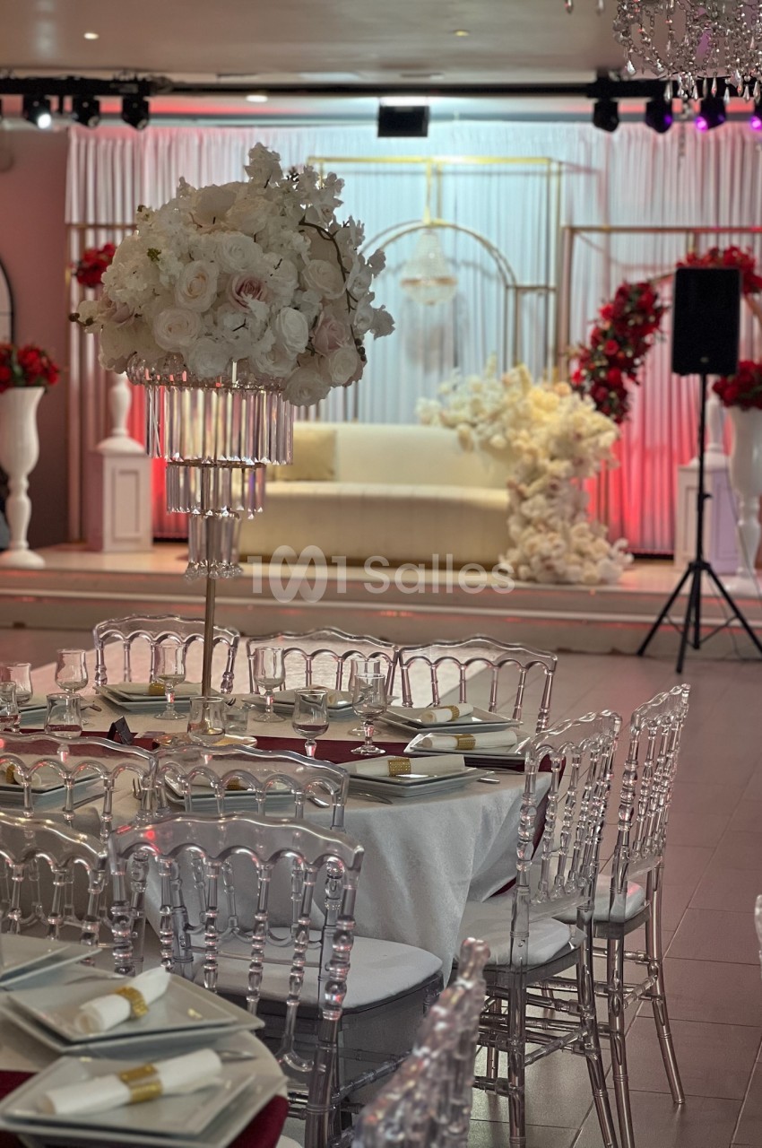 Salle décorée pour un événement avec tables dressées, chaises transparentes et arrangements floraux blancs et rouges.