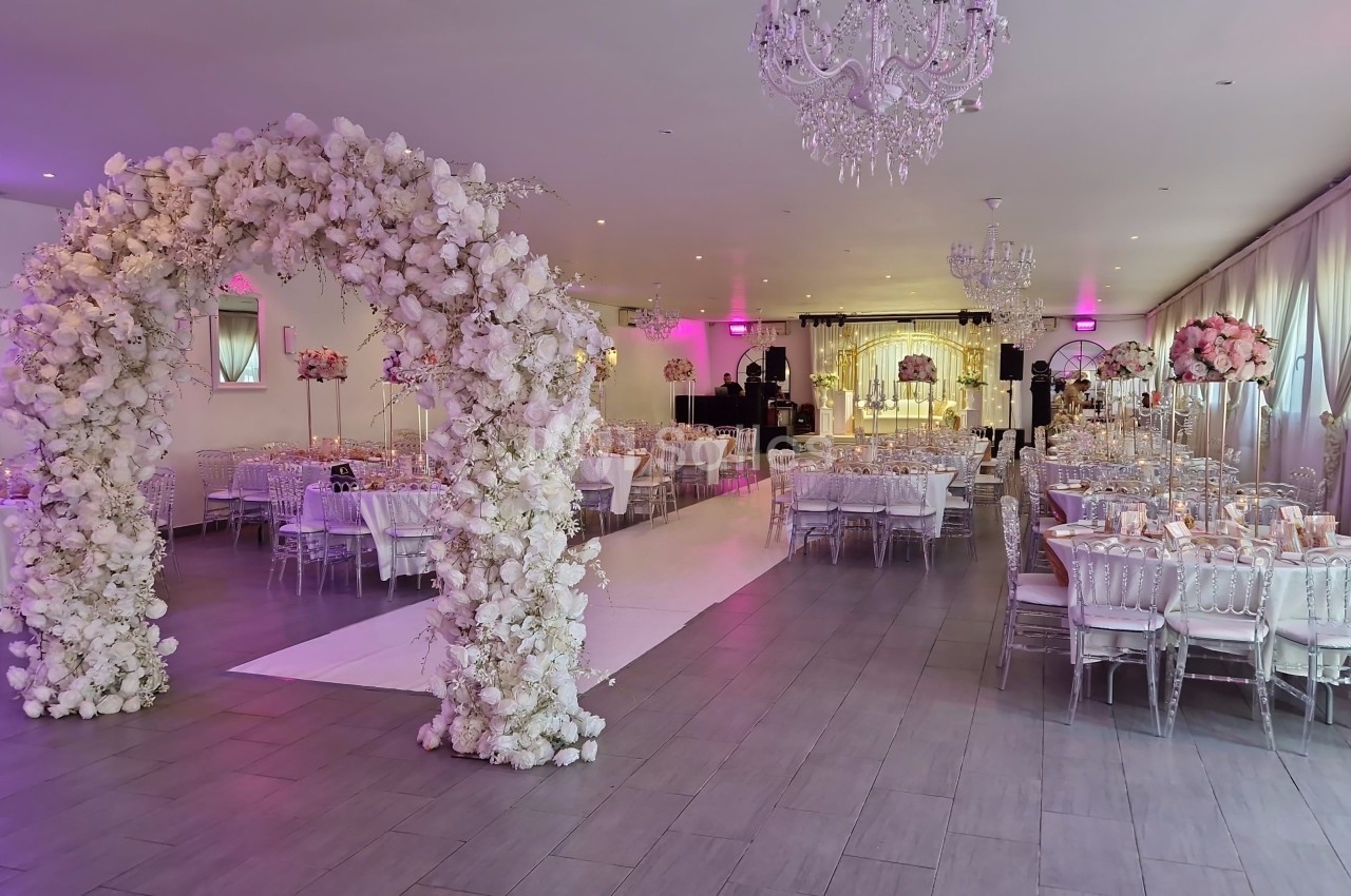 Salle de réception décorée avec des tables élégantes, un arche floral blanc et un éclairage rose tamisé.