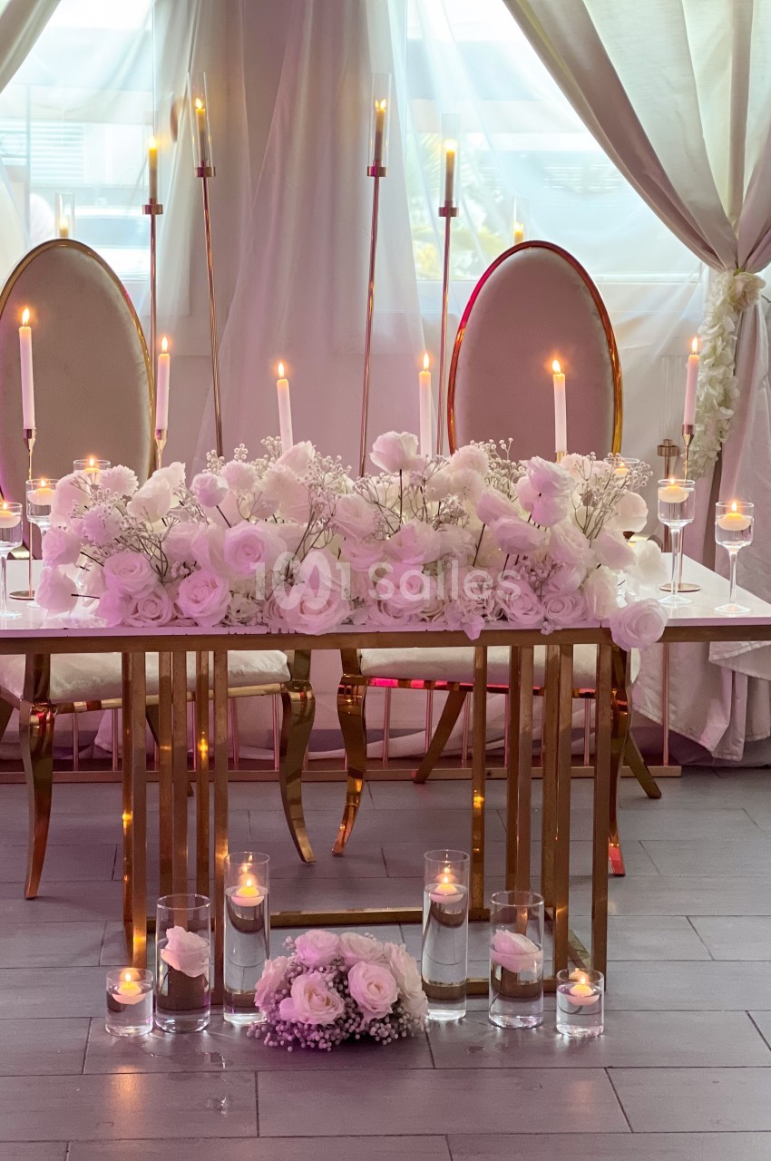 Table décorée avec des fleurs blanches et roses, entourée de bougies allumées et de chaises élégantes devant des rideaux.