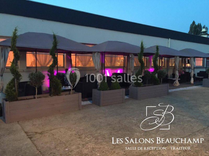 Terrasse aménagée avec des tentes, des plantes en bacs et un éclairage violet, devant un bâtiment moderne.