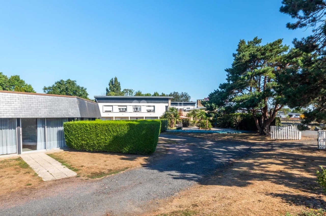 Vue d'un bâtiment moderne entouré d'arbres et de haies, avec une allée en gravier et une pelouse partiellement sèche.