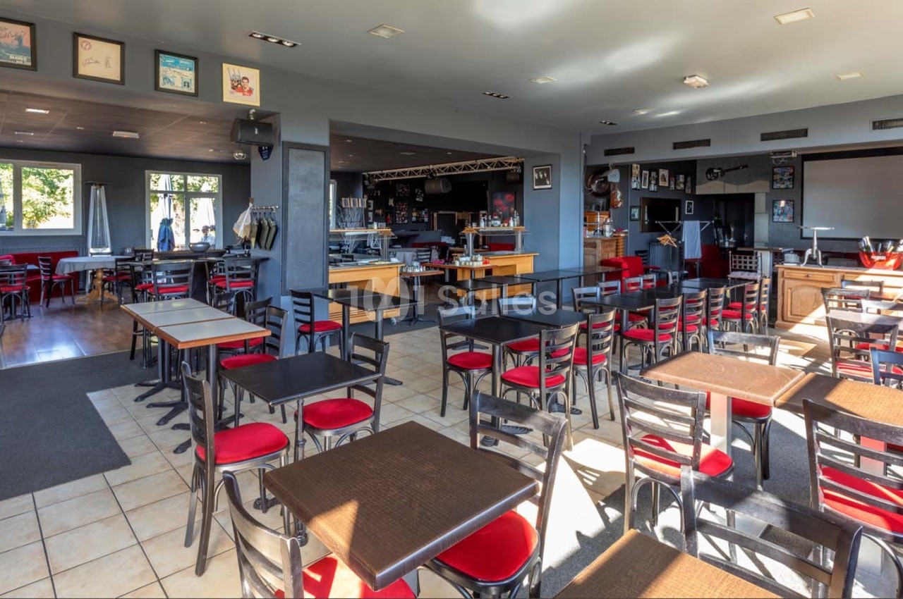 Salle de restaurant lumineuse avec des tables et chaises noires, assises rouges, et une scène en arrière-plan.