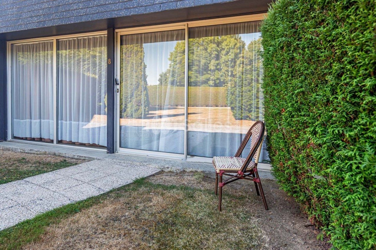Chaise en osier placée près d'une haie verte devant une maison avec de grandes baies vitrées et un chemin pavé.