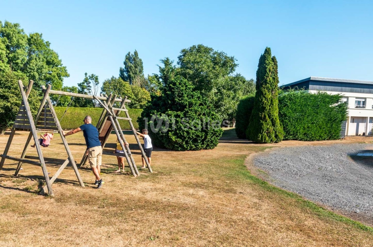 Un adulte et deux enfants jouent sur une balançoire en bois dans un espace extérieur avec arbres et pelouse.