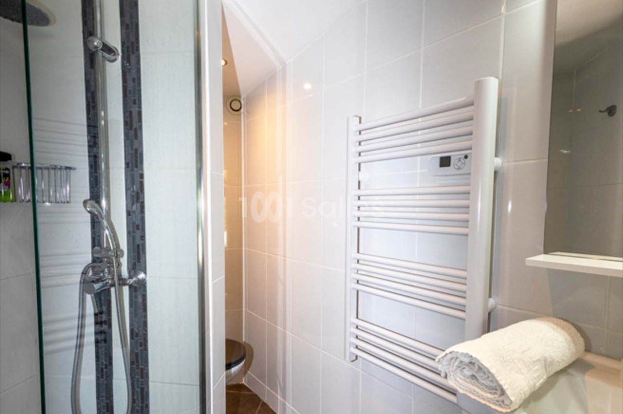 Salle de bain moderne avec douche, sèche-serviettes mural, carrelage blanc et serviette pliée sur une étagère.