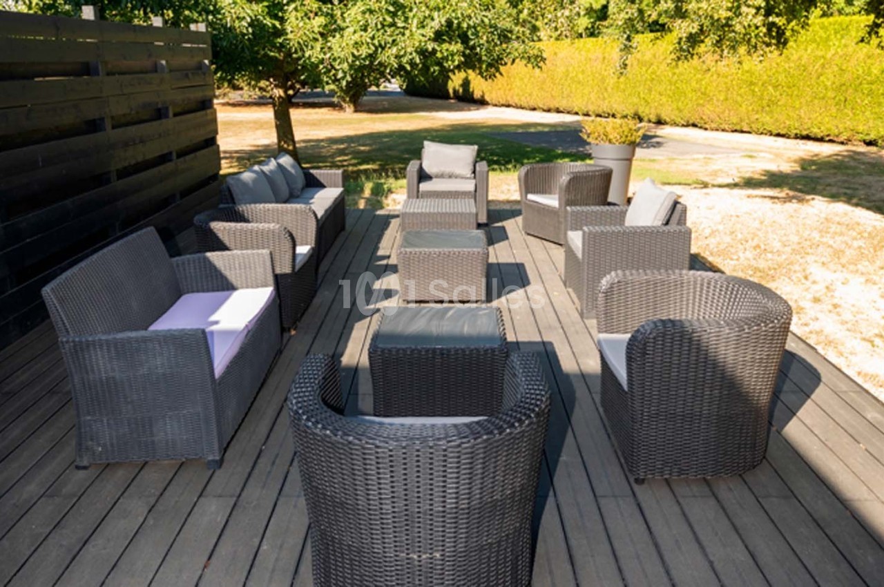 Salon de jardin en résine tressée avec fauteuils et canapés disposés en cercle sur une terrasse en bois, entourée de verdure.