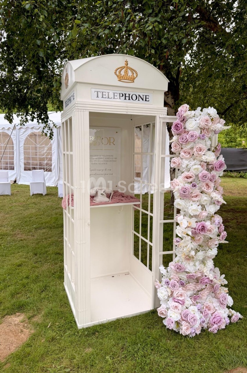 Cabine téléphonique blanche décorée de fleurs roses et blanches, installée sur une pelouse près de tentes blanches.