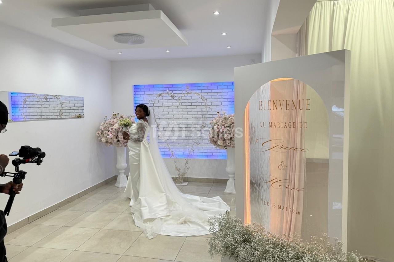 Une mariée en robe blanche pose devant une décoration florale et un panneau de bienvenue dans une salle lumineuse.