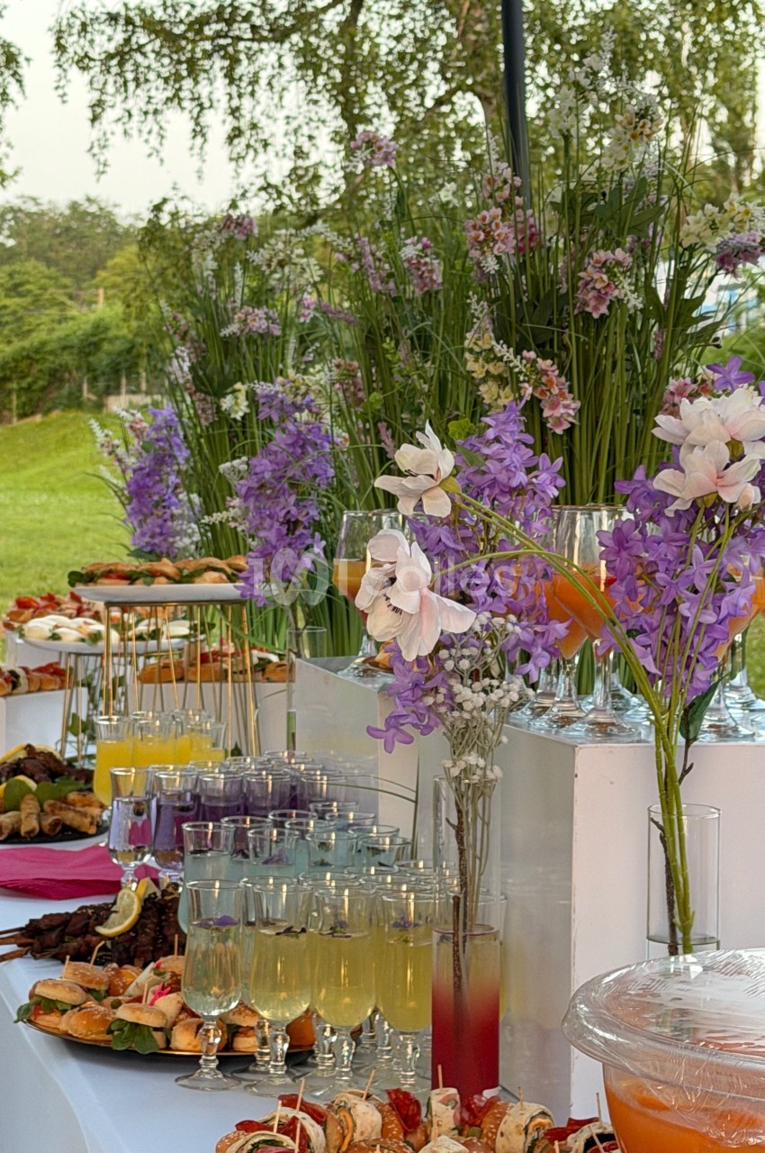 Buffet en plein air avec boissons colorées, brochettes, pâtisseries et décorations florales violettes et blanches.
