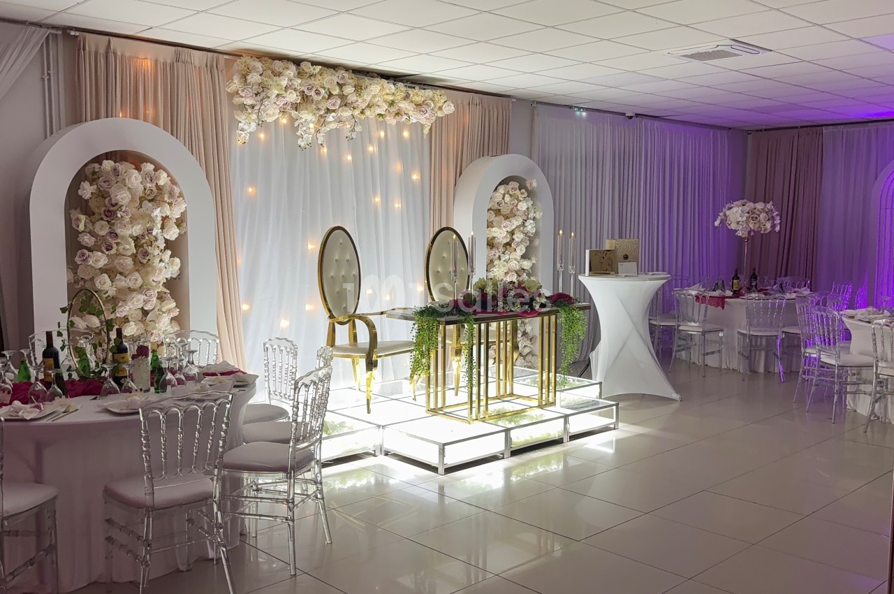 Salle de réception décorée avec des fleurs blanches, des chaises transparentes et une table d'honneur illuminée.