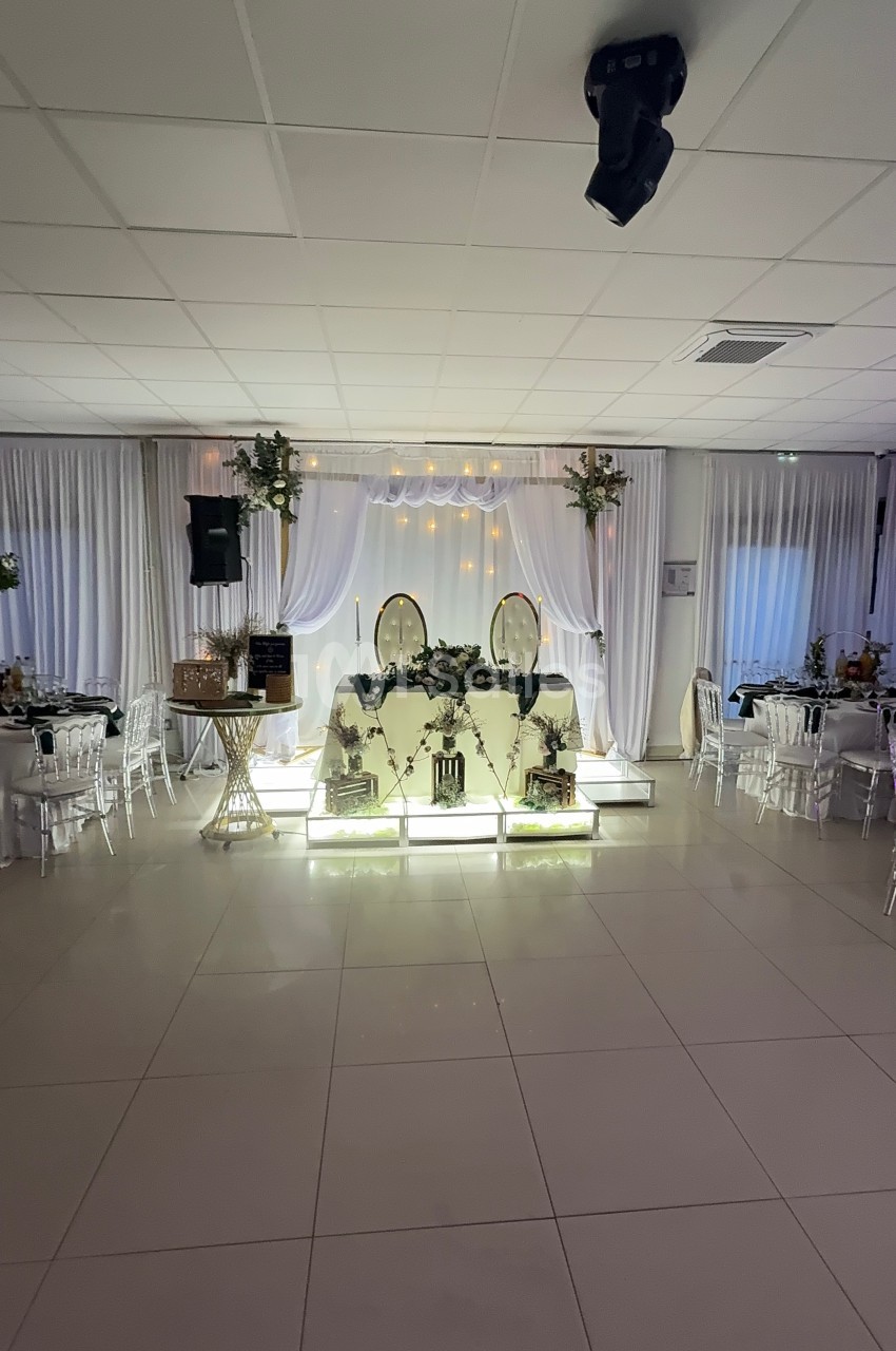 Salle de réception décorée avec une table d'honneur illuminée, ornée de fleurs et entourée de tables dressées.